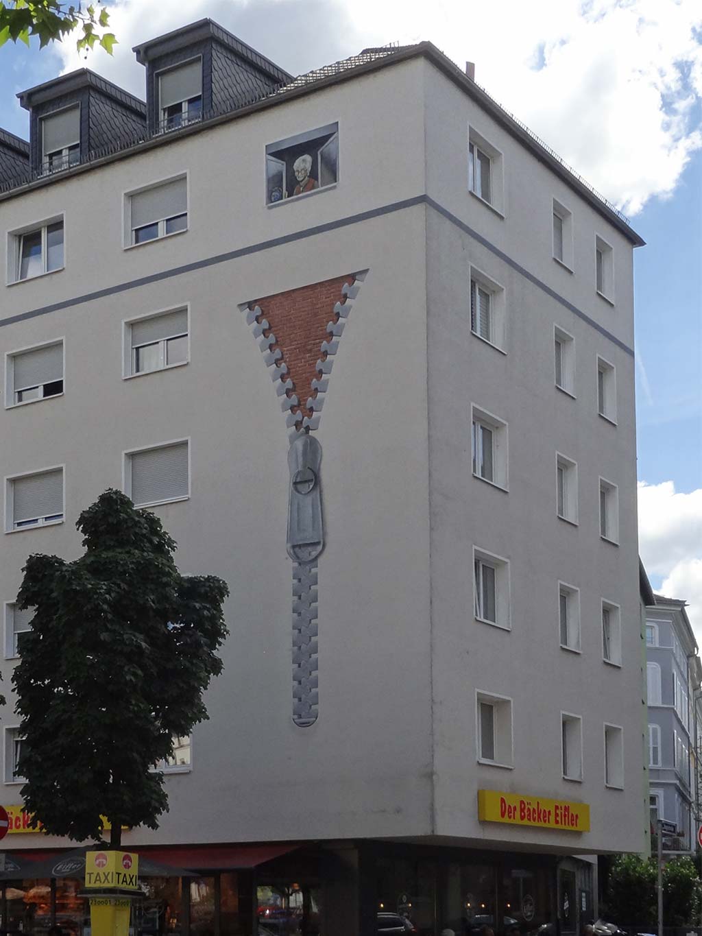 Geöffneter Reisverschluss gibt Blick frei auf Backsteinfassade, darüber schaut eine ältere Frau mit Bembel aus dem Fenster
